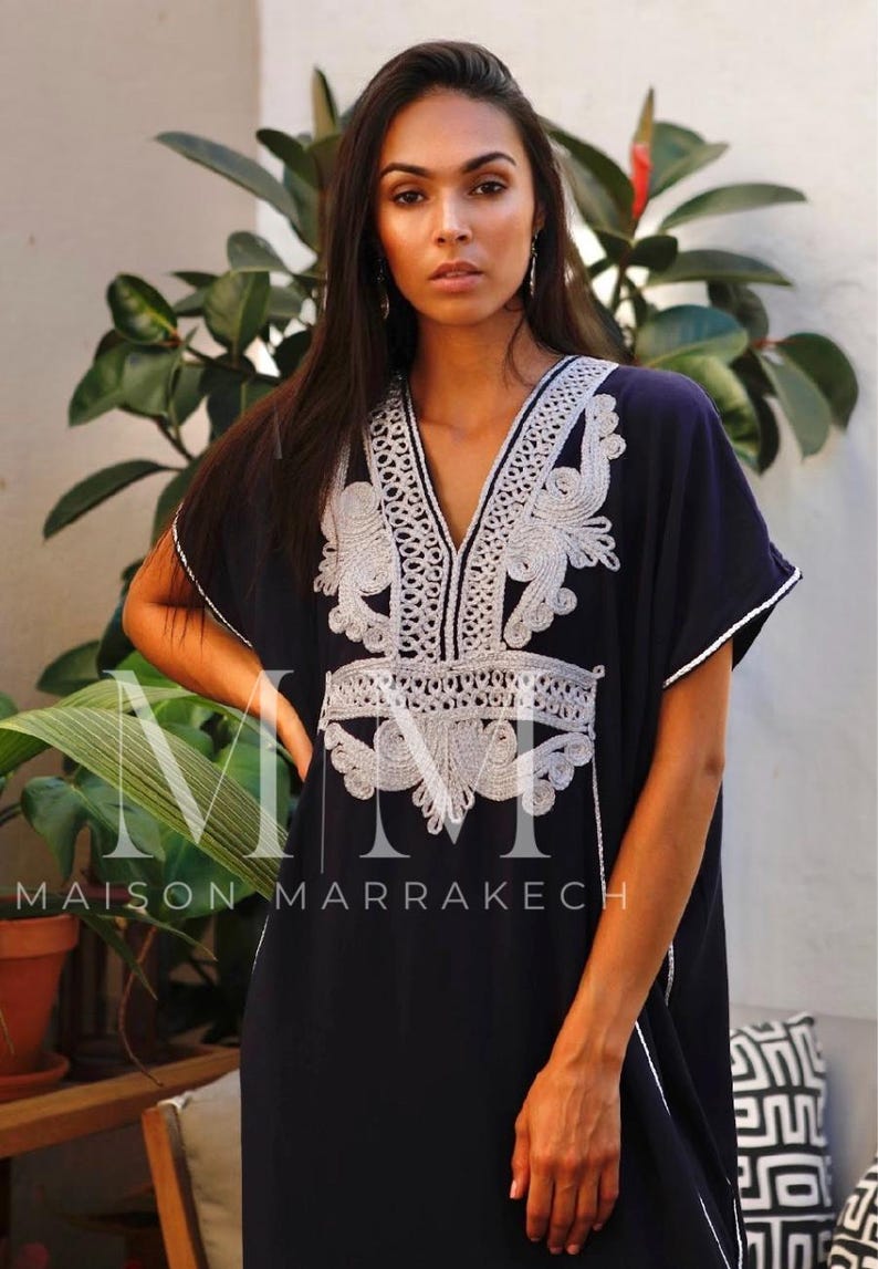 May include: A navy blue kaftan with white embroidery. The embroidery features a floral design and is accented with silver thread. The kaftan has a V-neckline and short sleeves. The text "MAISON MARRAKECH" is visible at the bottom of the image.