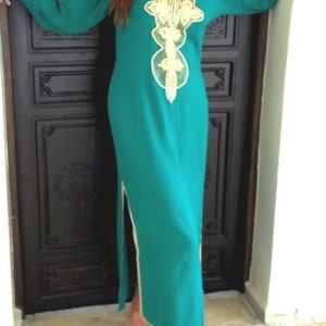 May include: A long, teal blue dress with gold embroidery and a V-neckline. The dress has a slit on the side and is worn by a person standing in front of a wooden door.