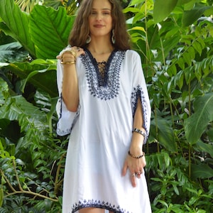 May include: A white tunic with blue embroidery and a lace-up neckline. The tunic has a loose fit and 3/4 sleeves. The model is wearing the tunic with a variety of gold rings and bracelets.