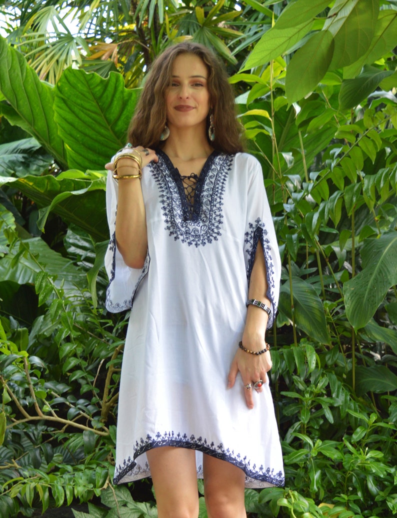 May include: A white tunic with blue embroidery and a lace-up neckline. The tunic has a loose fit and 3/4 sleeves. The model is wearing the tunic with a variety of gold rings and bracelets.