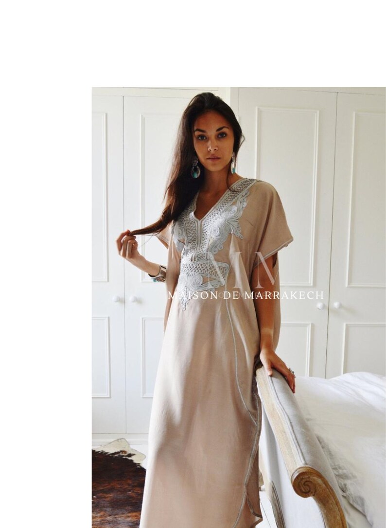 Puede incluir: Un vestido caft&aacute;n beige con intrincados bordados plateados alrededor del escote y las mangas. El vestido tiene un cuello en V y un dise&ntilde;o largo y fluido. El texto "MAISON DE MARRAKECH" es visible.