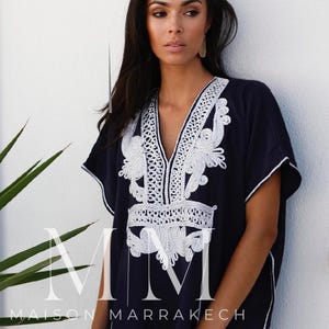 May include: A navy blue kaftan with white embroidery. The kaftan has a V-neckline and short sleeves. The embroidery is intricate and features floral designs. The text "Maison Marrakech" is visible on the bottom of the kaftan.