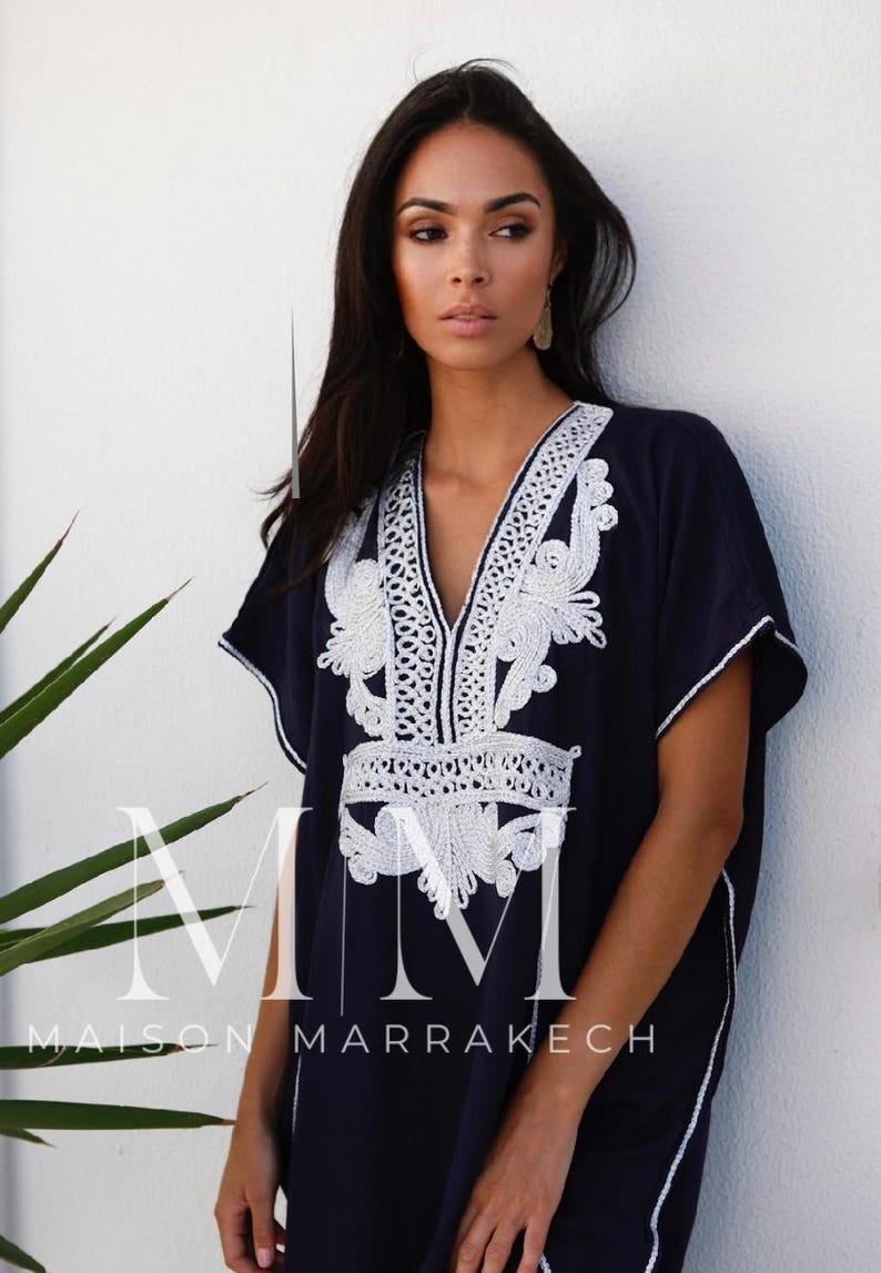 May include: A navy blue kaftan with white embroidery. The kaftan has a V-neckline and short sleeves. The embroidery is intricate and features floral designs. The text "Maison Marrakech" is visible on the bottom of the kaftan.