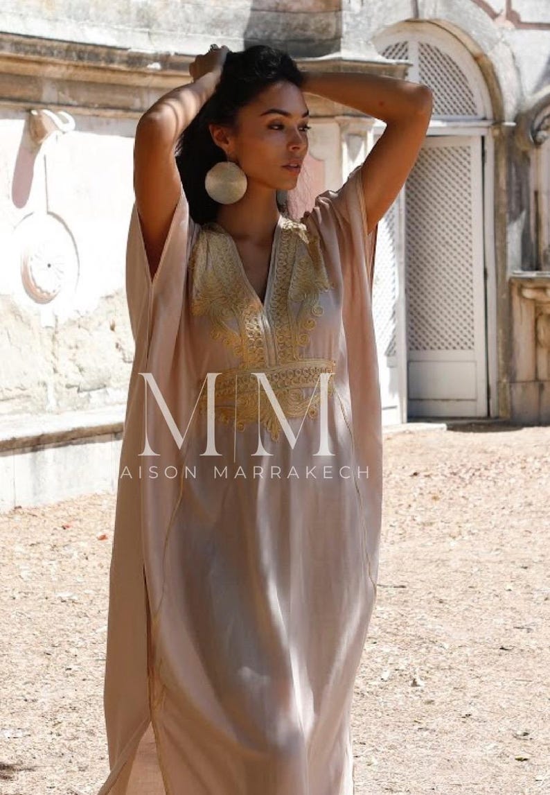 May include: A woman wearing a long, light pink kaftan with gold embroidery. The kaftan has a V-neck and a long, flowing skirt. The woman is standing in front of a building with a white door. The text "MM Maison Marrakech" is visible on the bottom of the image.
