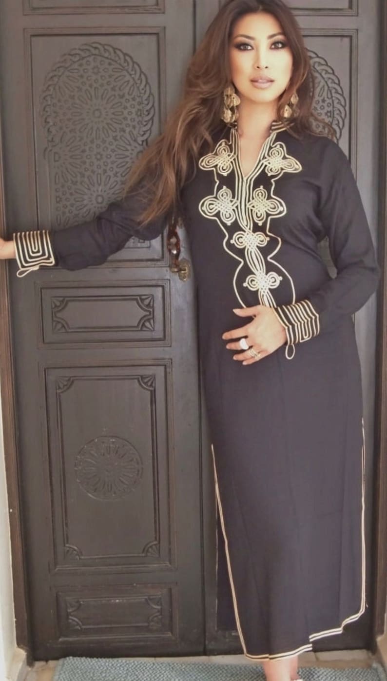 K&ouml;nnte beinhalten: Eine Frau tr&auml;gt ein schwarzes, lang&auml;rmeliges Kleid mit goldener Stickerei. Das Kleid hat einen V-Ausschnitt und einen langen Rock. Die Frau steht vor einer Holzt&uuml;r mit aufwendigen Schnitzereien.