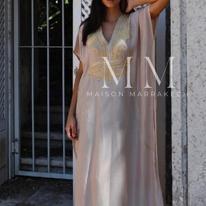 May include: A long, flowing, light beige kaftan with gold embroidery. The kaftan has a V-neck and short sleeves. The embroidery features a floral design and the text "MM MAISON MARRAKECH".