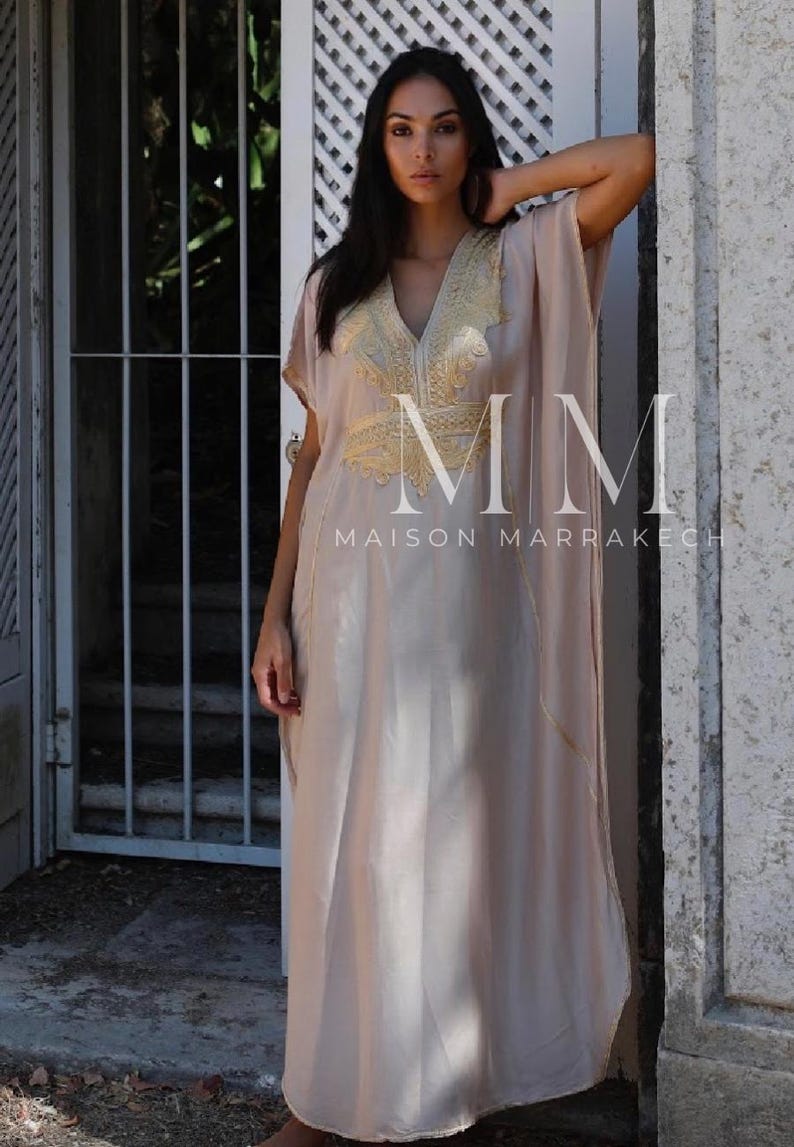 May include: A long, flowing, light beige kaftan with gold embroidery. The kaftan has a V-neck and short sleeves. The embroidery features a floral design and the text "MM MAISON MARRAKECH".