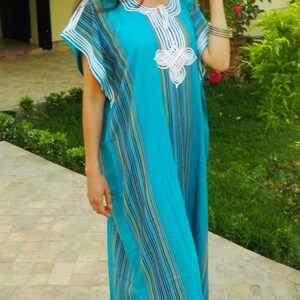 May include: A woman wearing a long, blue and white striped dress with white embroidery. The dress has a V-neck and a slit on the side. She is standing on a tiled floor with green plants in the background.
