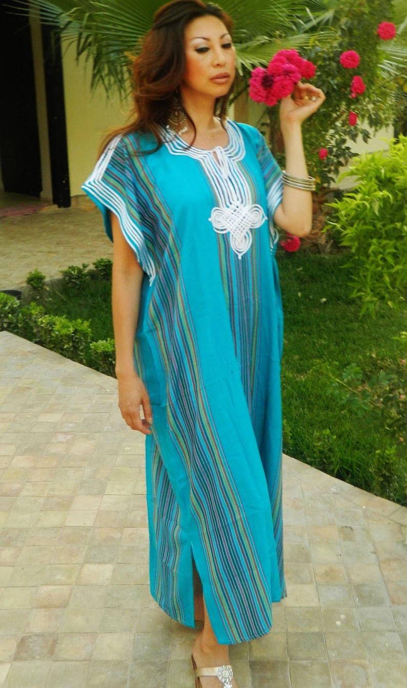 May include: A woman wearing a long, blue and white striped dress with white embroidery. The dress has a V-neck and a slit on the side. She is standing on a tiled floor with green plants in the background.