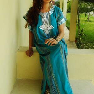 May include: A woman wearing a long, turquoise blue and white striped dress with white embroidery. She is sitting on a step with her legs crossed and wearing silver sandals.