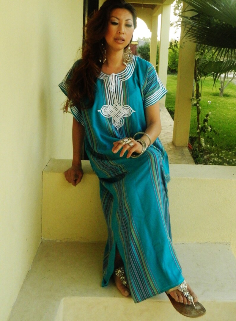 May include: A woman wearing a long, turquoise blue and white striped dress with white embroidery. She is sitting on a step with her legs crossed and wearing silver sandals.