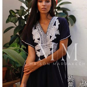 May include: A black kaftan with white embroidery and a V-neckline. The kaftan has short sleeves and a long, flowing skirt. The text "Maison Marrakech" is printed on the front of the kaftan.