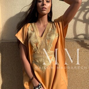 May include: A long, loose-fitting, orange kaftan with gold embroidery. The kaftan has a V-neck and short sleeves. The text "Maison Marrakech" is visible on the bottom of the garment.