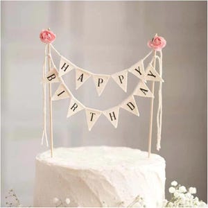 May include: White cake with a happy birthday banner made of fabric flags with pink flowers on each end. The banner says "Happy Birthday".