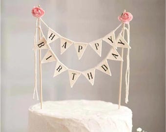 May include: White cake with a happy birthday banner made of fabric flags with pink flowers on each end. The banner says "Happy Birthday".