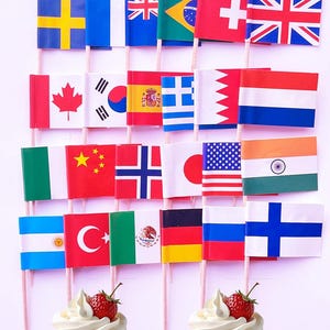 以下が含まれることがあります： アメリカ、カナダ、メキシコ、ドイツ、フランスなど、さまざまな国を代表する木製スティックに20本のミニチュアフラッグがセットされています。フラッグは白い背景に並べて配置され、ホイップクリームとイチゴをトッピングしたカップケーキが2つ前景にあります。