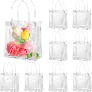 Puede incluir: Bolso tote de plástico transparente con un ramo de flores rosas y amarillas. El bolso tiene asas transparentes y está rodeado de varios otros bolsos transparentes vacíos.