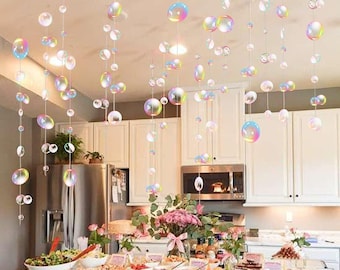 Puede incluir: Una cocina decorada para una fiesta con una guirnalda de burbujas iridiscentes colgando del techo. La guirnalda está hecha de burbujas de plástico transparente con un acabado iridiscente arcoíris. Las burbujas están enhebradas en una línea de pesca transparente y cuelgan a diferentes longitudes. Hay una mesa en primer plano con comida y bebidas sobre ella.
