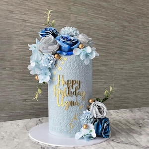 Puede incluir: Una tarta de cumpleaños azul claro con letras doradas que dicen "Happy Birthday Uzma". La tarta está decorada con flores azules, grises y blancas y detalles dorados. La tarta está sobre una base blanca.