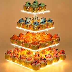 Può includere: Un porta cupcake in acrilico a quattro livelli con pirottini per cupcake con bordo dorato. Il porta cupcake è illuminato da luci LED bianco caldo e presenta una varietà di cupcake colorati.