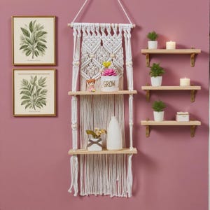 May include: A macramé hanging shelf unit with three wooden shelves, decorated with small plants, candles, and ornaments. Two framed botanical prints are on the wall. The shelf is suspended from a wooden dowel.