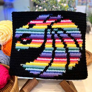 May include: A rainbow crochet lion head design on a black background. The crochet piece is displayed on a wooden easel.