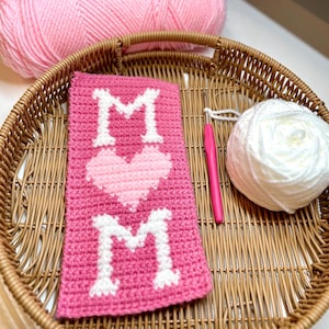 May include: A woven basket holds a pink crocheted item with the word "MOM" and a heart design, a pink crochet hook, and a ball of white yarn. Two pink yarn skeins are in the background.