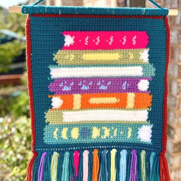 Peut inclure: Une tenture murale crochetée à la main représentant une pile de livres colorés. Les livres sont dans les tons de rose, jaune, blanc, violet, orange et bleu. La tenture a un fond turquoise avec une bordure et des franges rouges.