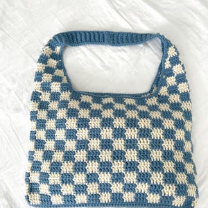 Peut inclure: Un sac fourre-tout crochet&eacute; bleu et cr&egrave;me avec un motif &agrave; carreaux. Le sac a une forme rectangulaire avec une seule poign&eacute;e. Les points de crochet sont visibles et le sac semble &ecirc;tre fait &agrave; la main.
