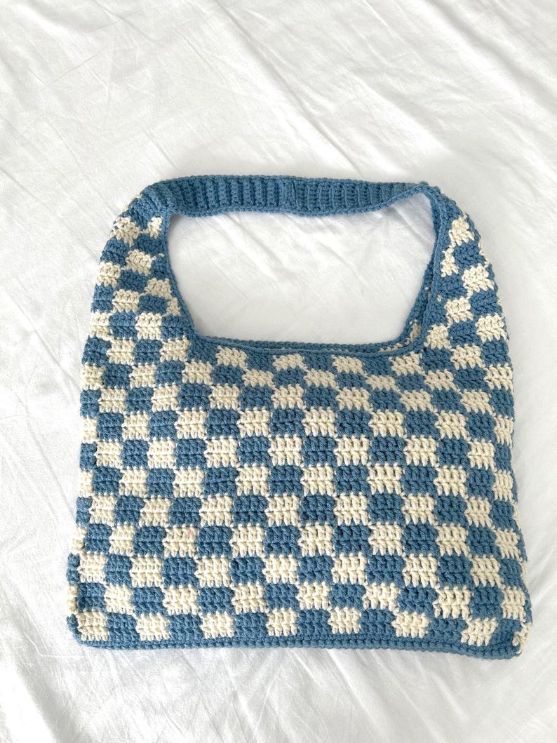 Peut inclure: Un sac fourre-tout crochet&eacute; bleu et cr&egrave;me avec un motif &agrave; carreaux. Le sac a une forme rectangulaire avec une seule poign&eacute;e. Les points de crochet sont visibles et le sac semble &ecirc;tre fait &agrave; la main.