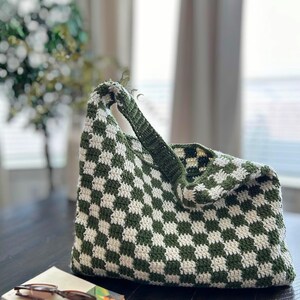 Peut inclure: Un sac fourre-tout crochet&eacute; &agrave; la main, motif damier vert et blanc. Le sac a une seule poign&eacute;e et est pos&eacute; sur une surface en bois fonc&eacute;. Un livre et des lunettes sont &agrave; c&ocirc;t&eacute; du sac.