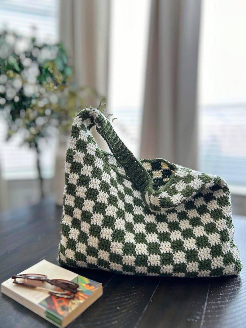 Peut inclure: Un sac fourre-tout crochet&eacute; &agrave; la main, motif damier vert et blanc. Le sac a une seule poign&eacute;e et est pos&eacute; sur une surface en bois fonc&eacute;. Un livre et des lunettes sont &agrave; c&ocirc;t&eacute; du sac.