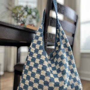 Peut inclure: Un sac fourre-tout en crochet fait main, motif damier bleu et cr&egrave;me. Il est suspendu &agrave; une chaise en bois. Le sac a de longues anses et une ouverture r&eacute;v&eacute;lant un int&eacute;rieur spacieux, id&eacute;al pour transporter des effets personnels.