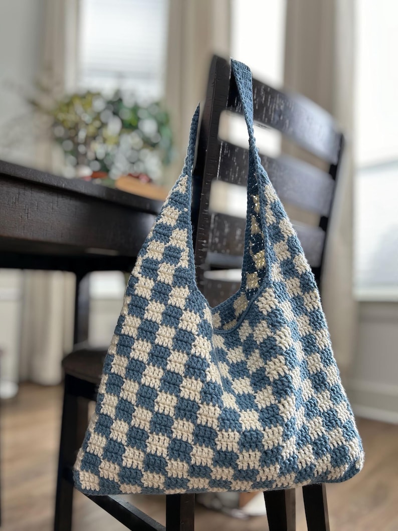 Peut inclure: Un sac fourre-tout en crochet fait main, motif damier bleu et cr&egrave;me. Il est suspendu &agrave; une chaise en bois. Le sac a de longues anses et une ouverture r&eacute;v&eacute;lant un int&eacute;rieur spacieux, id&eacute;al pour transporter des effets personnels.