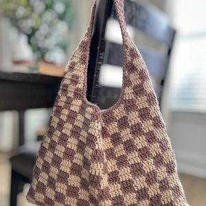 Peut inclure: Un sac fourre-tout crochet&eacute; &agrave; la main avec un motif &agrave; carreaux mauve et cr&egrave;me. Le sac a un dessus large et ouvert et de longues sangles pour le porter &agrave; l'&eacute;paule. Le sac est accroch&eacute; au dossier d'une chaise en bois fonc&eacute;.