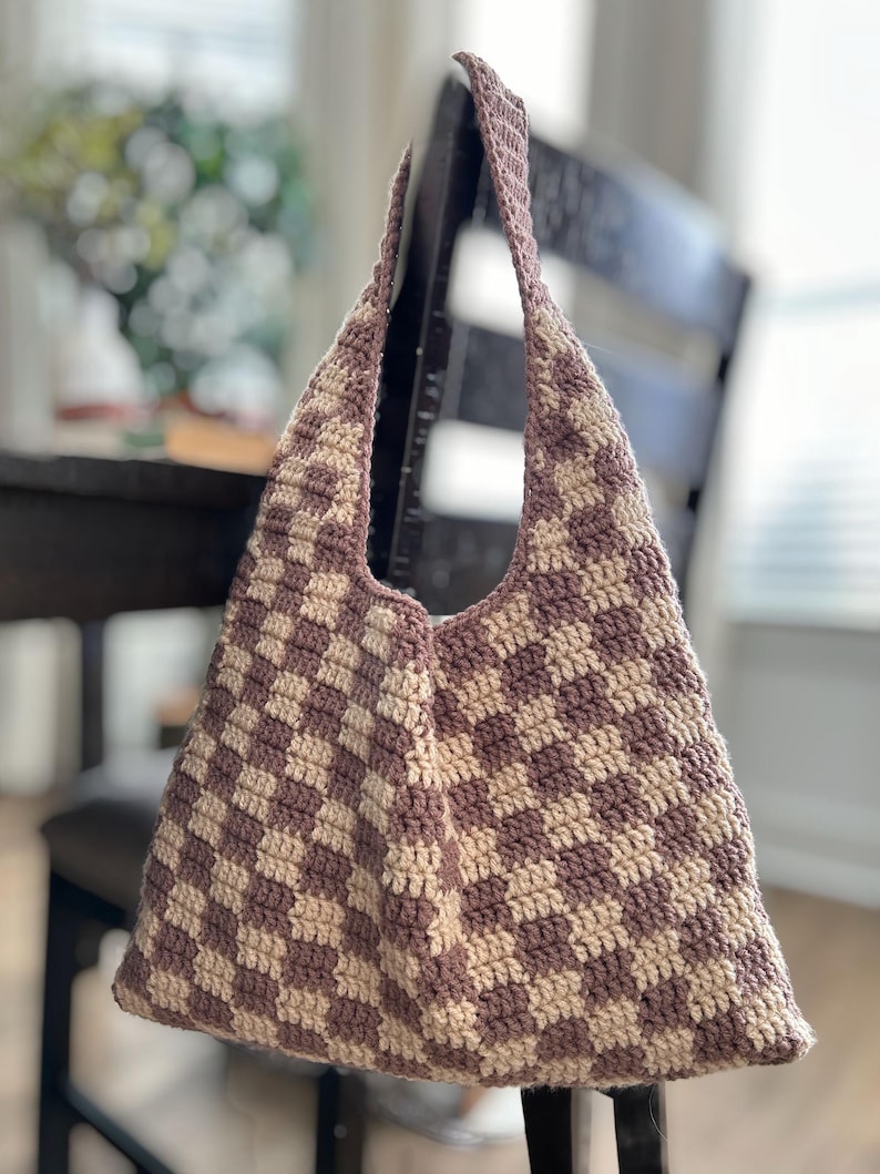 Peut inclure: Un sac fourre-tout crochet&eacute; &agrave; la main avec un motif &agrave; carreaux mauve et cr&egrave;me. Le sac a un dessus large et ouvert et de longues sangles pour le porter &agrave; l'&eacute;paule. Le sac est accroch&eacute; au dossier d'une chaise en bois fonc&eacute;.