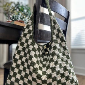 Peut inclure: Un sac fourre-tout crochet&eacute; &agrave; la main avec un motif &agrave; carreaux vert et blanc. Le sac a une large bandouli&egrave;re et est suspendu &agrave; une chaise en bois fonc&eacute;. Le design du sac est un motif g&eacute;om&eacute;trique classique.