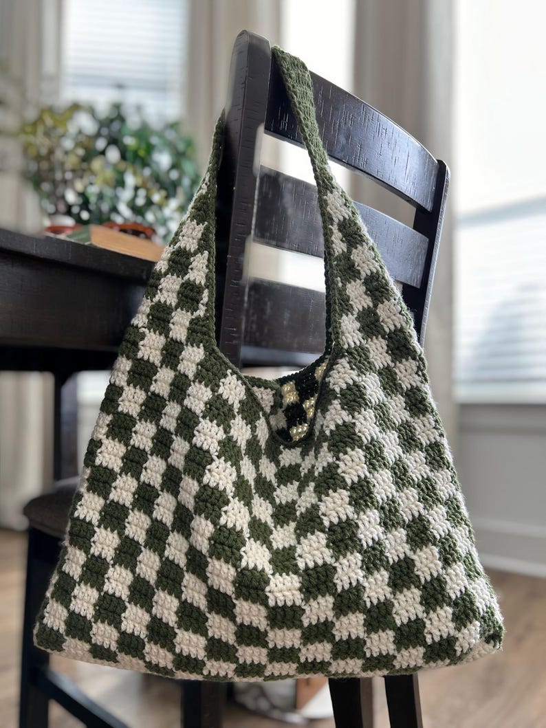 Peut inclure: Un sac fourre-tout crochet&eacute; &agrave; la main avec un motif &agrave; carreaux vert et blanc. Le sac a une large bandouli&egrave;re et est suspendu &agrave; une chaise en bois fonc&eacute;. Le design du sac est un motif g&eacute;om&eacute;trique classique.
