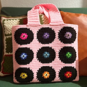 May include: A handmade pink crochet tote bag featuring nine black vinyl record designs with colorful center labels. The bag has a matching pink handle and is displayed on a green surface with decorative pillows in the background.