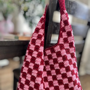 Peut inclure: Un sac fourre-tout crochet&eacute; &agrave; la main avec un motif en damier rouge fonc&eacute; et rose clair. Le sac est suspendu &agrave; une chaise en bois fonc&eacute;. La sangle et le bord sup&eacute;rieur du sac sont d'un rouge fonc&eacute; uni.