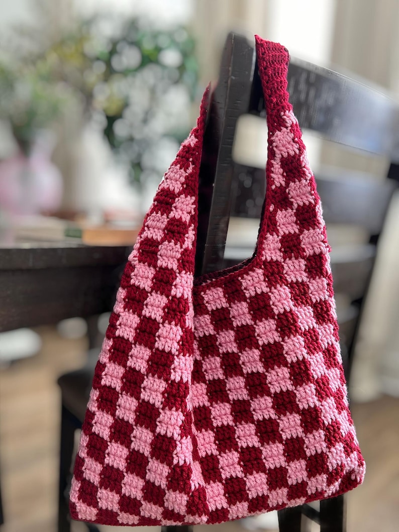 Peut inclure: Un sac fourre-tout crochet&eacute; &agrave; la main avec un motif en damier rouge fonc&eacute; et rose clair. Le sac est suspendu &agrave; une chaise en bois fonc&eacute;. La sangle et le bord sup&eacute;rieur du sac sont d'un rouge fonc&eacute; uni.