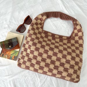 Peut inclure: Un sac fourre-tout en crochet fait &agrave; la main avec un motif &agrave; carreaux marron et beige. Le sac a une large poign&eacute;e et est pr&eacute;sent&eacute; avec des lunettes de soleil marron, un petit portefeuille et un livre sur une surface blanche.