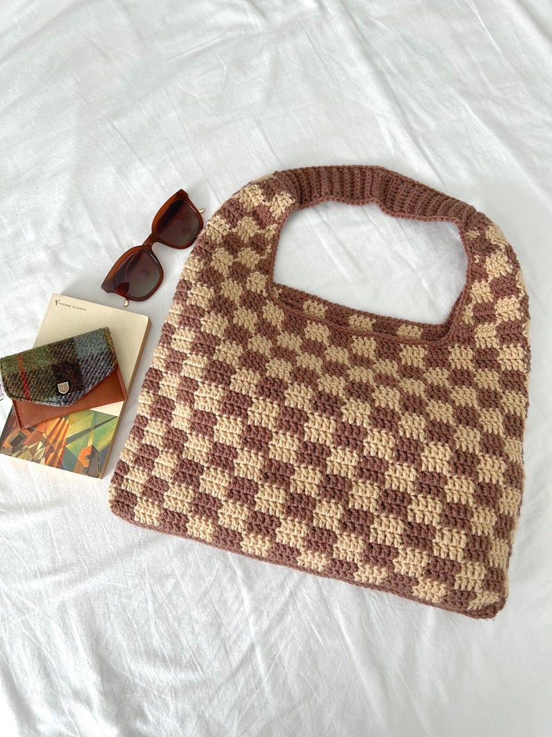 Peut inclure: Un sac fourre-tout en crochet fait &agrave; la main avec un motif &agrave; carreaux marron et beige. Le sac a une large poign&eacute;e et est pr&eacute;sent&eacute; avec des lunettes de soleil marron, un petit portefeuille et un livre sur une surface blanche.