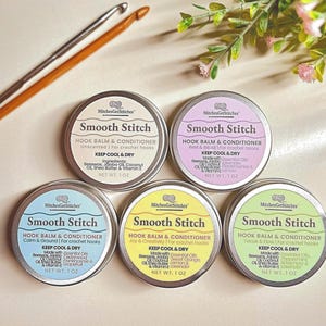 May include: An overhead shot displays five circular tins of "Smooth Stitch" hook balm and conditioner, each with a different color label. Two wooden crochet hooks are in the upper left. The tins are labeled with the product name and purpose.