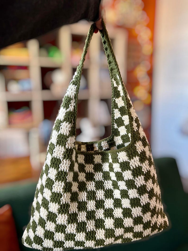 Peut inclure: Sac fourre-tout au crochet fait main avec un motif &agrave; carreaux vert et blanc. Le sac a une large ouverture et de longues sangles. Le sac est tenu par une main.