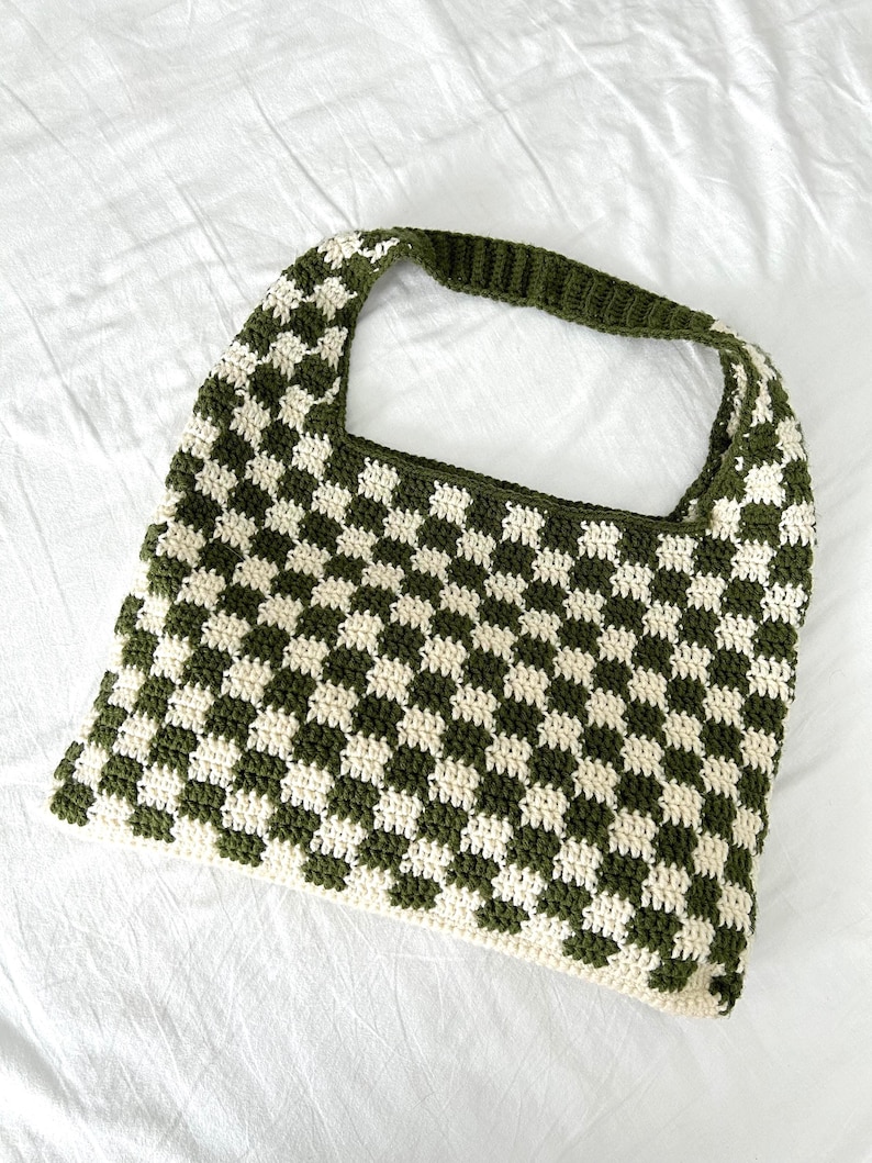 Peut inclure: Un sac fourre-tout crochet&eacute; &agrave; la main avec un motif en damier. Le sac pr&eacute;sente des carr&eacute;s altern&eacute;s vert olive et cr&egrave;me. La poign&eacute;e est de couleur vert olive uni. Le sac est fait d'un fil doux et textur&eacute;.