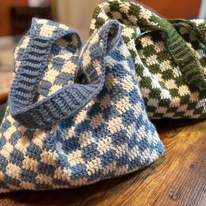 Peut inclure: Deux sacs fourre-tout crochet&eacute;s &agrave; la main. L'un est &agrave; carreaux bleus et blancs, avec une sangle bleue assortie. L'autre est &agrave; carreaux verts et blancs, avec une sangle verte. Les deux sacs sont pos&eacute;s sur une surface en bois.