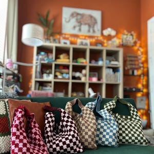 Peut inclure: Cinq sacs fourre-tout tricot&eacute;s &agrave; motif de damier, de couleurs rose, bordeaux, beige, bleu et vert. Les sacs sont faits &agrave; la main et sont pr&eacute;sent&eacute;s sur un canap&eacute; vert.