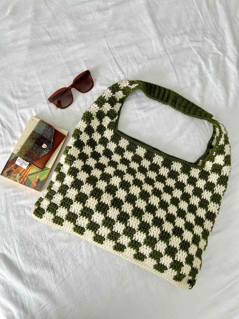 Peut inclure: Un sac fourre-tout crochet&eacute; &agrave; la main avec un motif &agrave; carreaux vert et cr&egrave;me. Le sac a une large bandouli&egrave;re. Sont &eacute;galement pr&eacute;sent&eacute;es des lunettes de soleil marron et un petit portefeuille avec un livre.