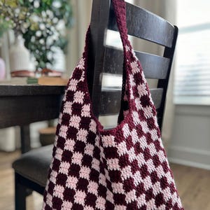 Peut inclure: Un sac fourre-tout crochet&eacute; &agrave; la main avec un motif en damier bordeaux et rose clair. Le sac est suspendu &agrave; une chaise en bois fonc&eacute;. La bandouli&egrave;re et le bord sup&eacute;rieur du sac sont bordeaux.